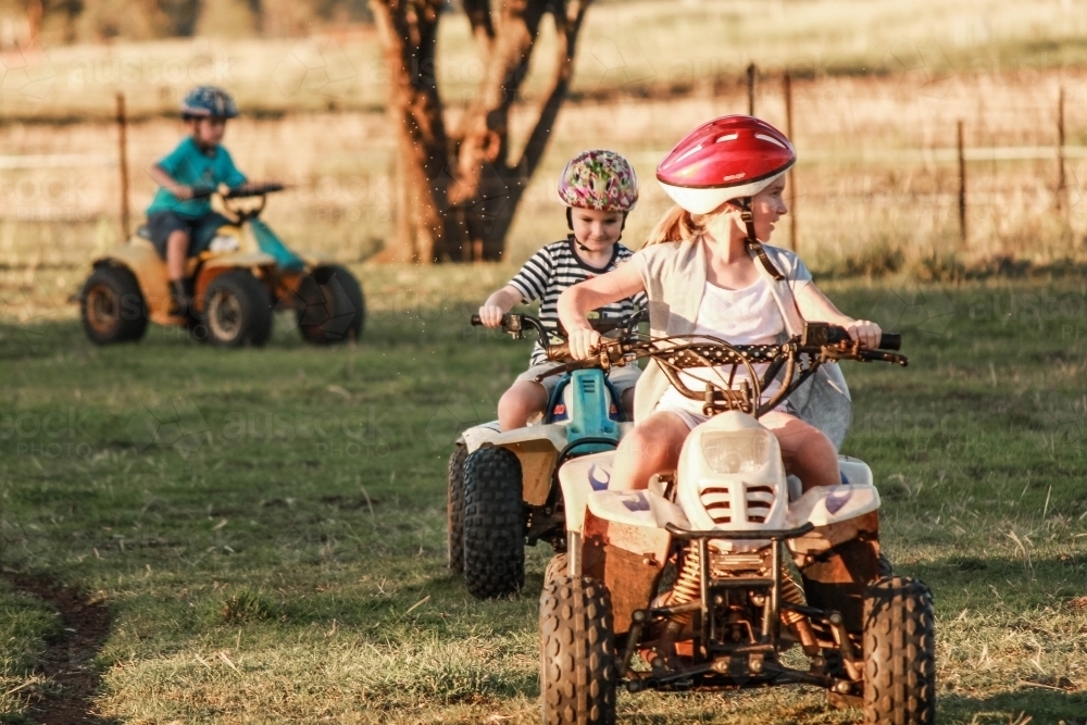Three children riding on quad bikes around farm property - Australian Stock Image