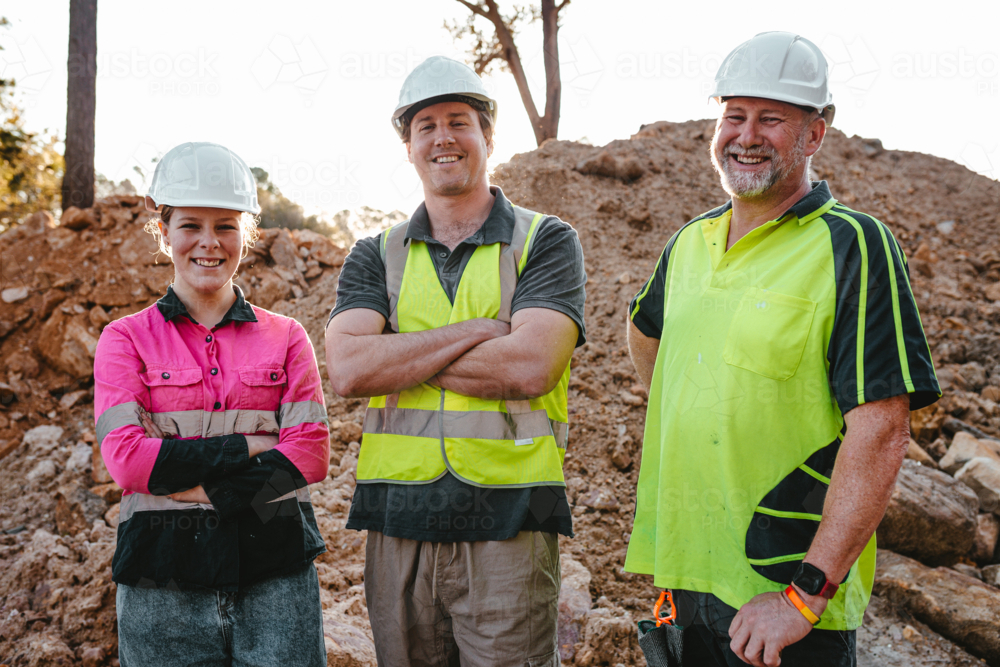 Three construction workers standing wearing safety equipment on the worksite with arms crossed - Australian Stock Image
