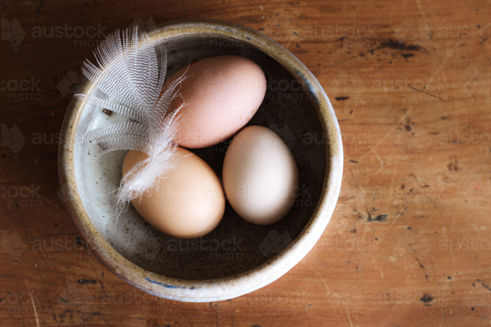 Three eggs with white feather on top in a rustic ceramic bowl sits on a wooden surface - Australian Stock Image