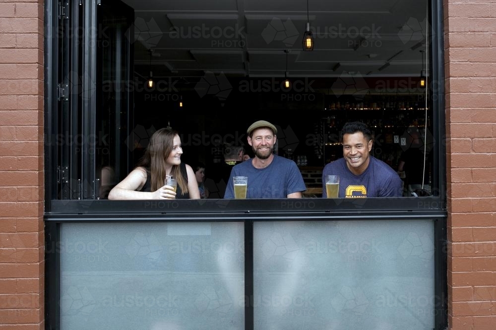 Three friends at a window having a drink at a local craft beer bar - Australian Stock Image