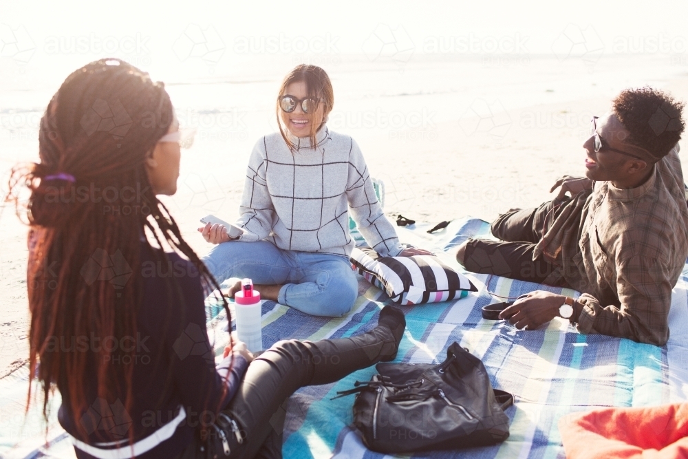 Three friends sitting on a picnic blanket relaxing at the beach - Australian Stock Image