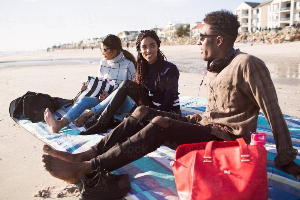 Three friends sitting on a picnic blanket relaxing at the beach - Australian Stock Image