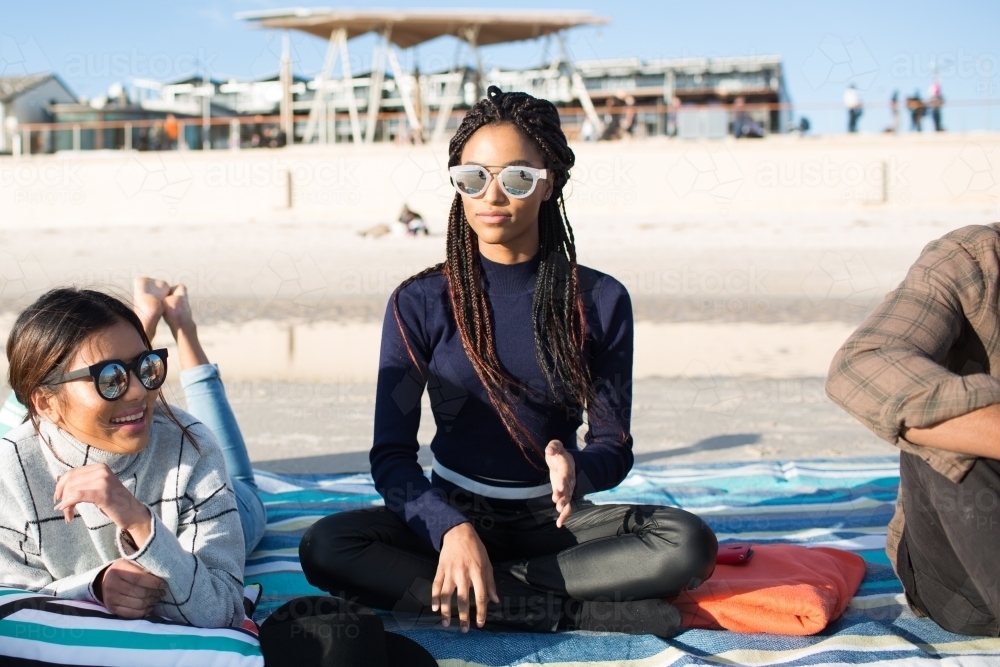 Three friends sitting on a picnic blanket relaxing at the beach - Australian Stock Image
