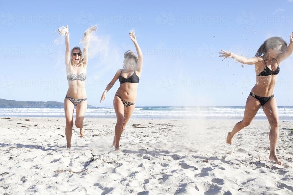 Three girls running on the beach throwing sand in the air - Australian Stock Image