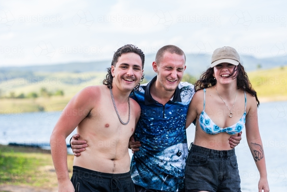 Three happy Aussie teenagers laughing together by lake in summer - Australian Stock Image