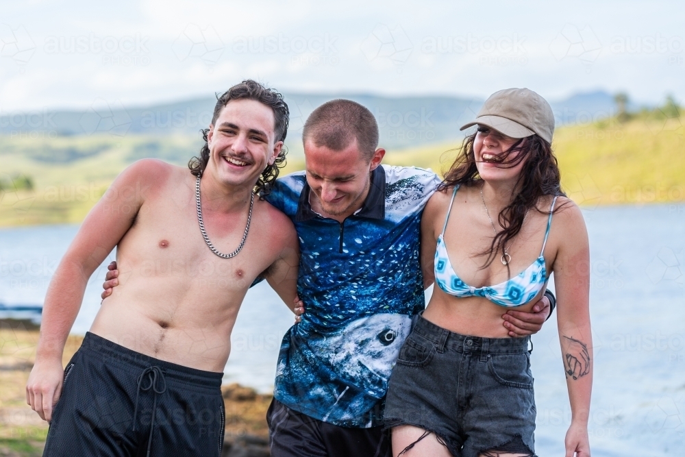 Three happy Aussie teenagers laughing together by lake in summer - Australian Stock Image