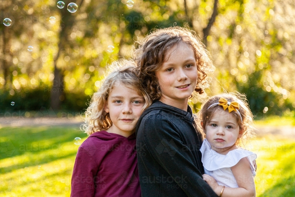 Three happy siblings with curly hair smiling in park - Australian Stock Image