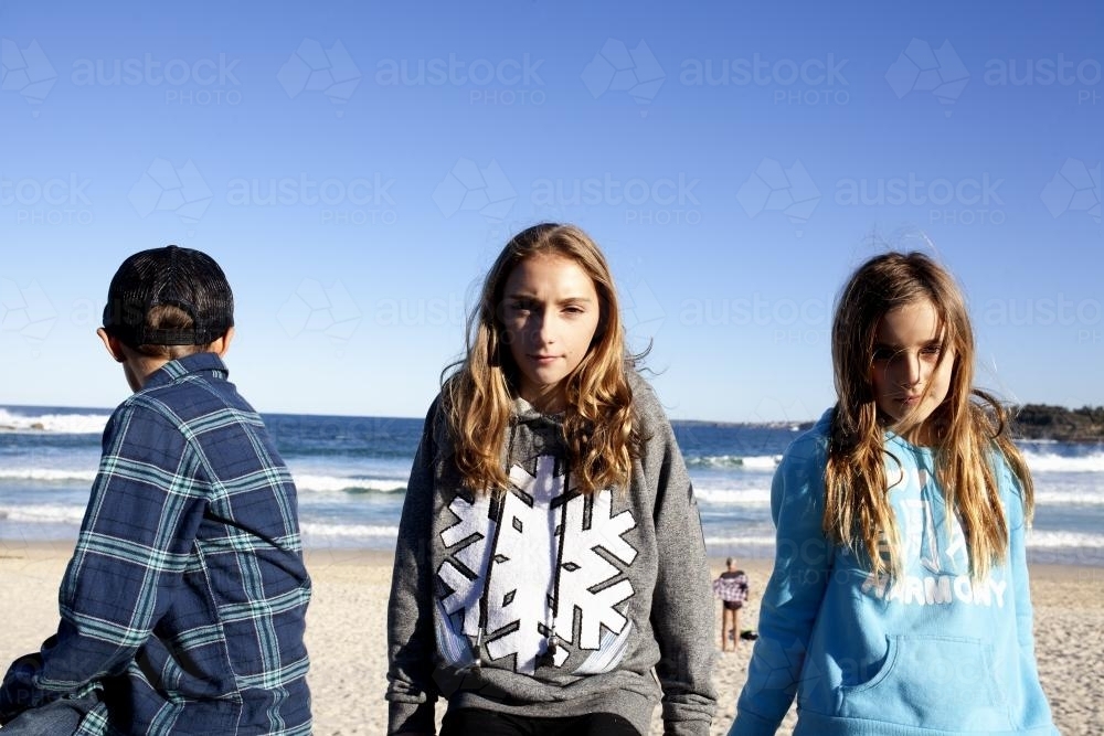 Three kids sitting in the sunshine on a railing in front of the ocean - Australian Stock Image