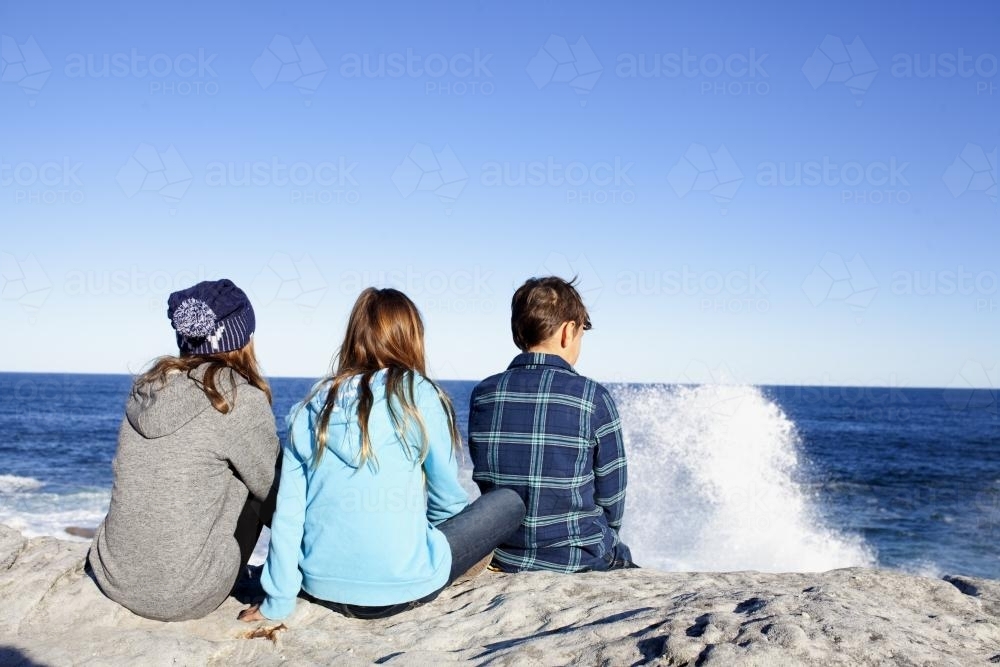 Three kids sitting on a rock looking out at the ocean - Australian Stock Image