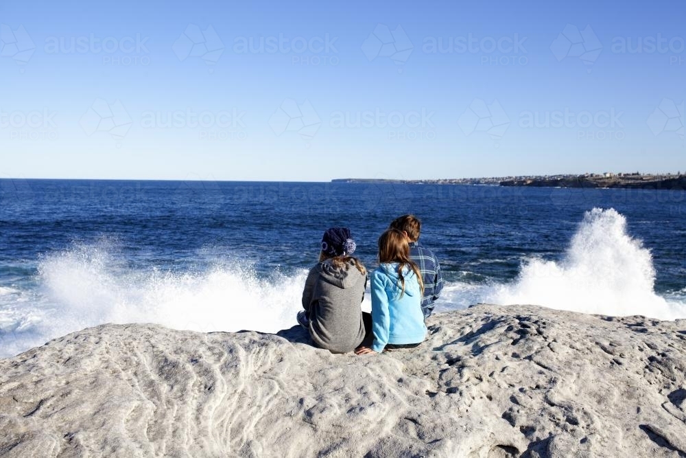 Three kids sitting on a rock looking out at the ocean with waves crashing - Australian Stock Image