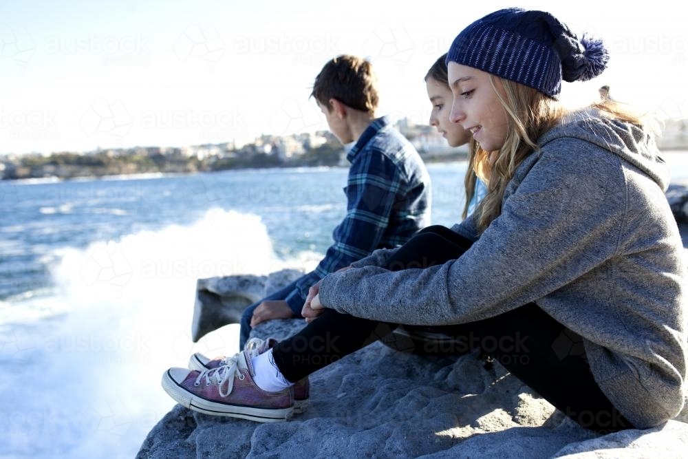 Three kids sitting on a rock lookout watching waves crash - Australian Stock Image