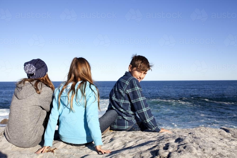 Three kids sitting on rock looking out at the ocean with boy looking behind him - Australian Stock Image