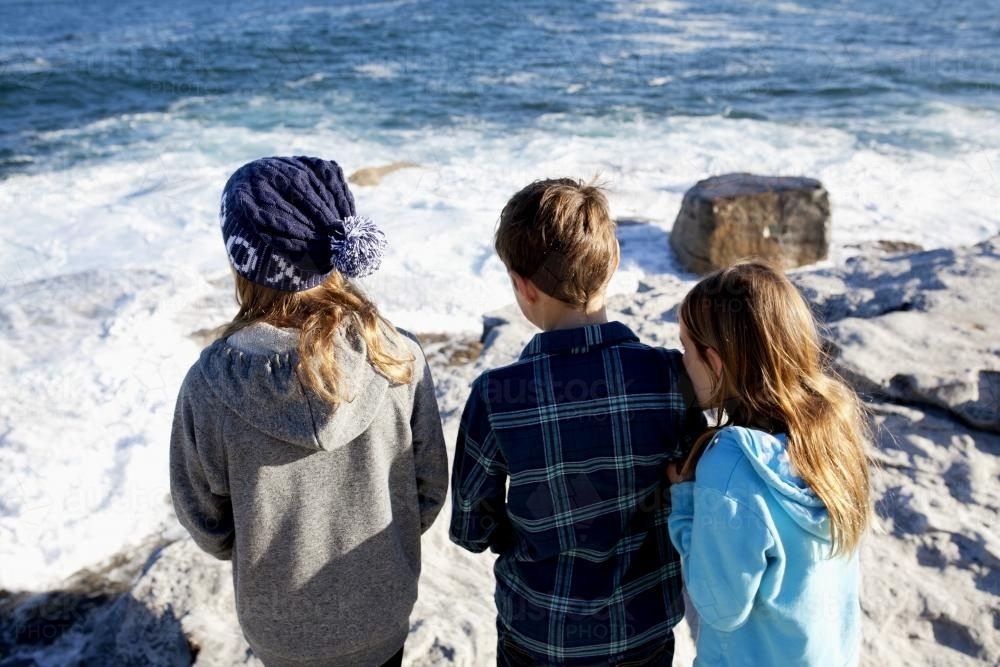 Three kids standing on a rock looking out at the ocean - Australian Stock Image