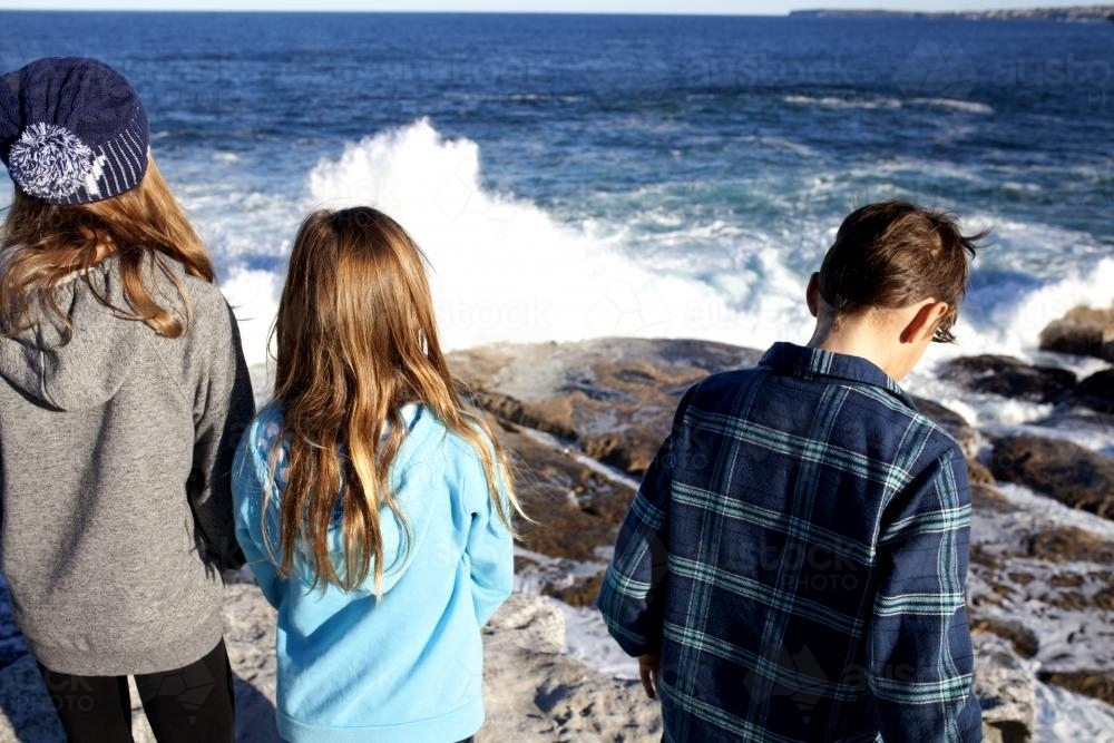 Three kids standing on a rock looking out at the ocean with waves crashing - Australian Stock Image