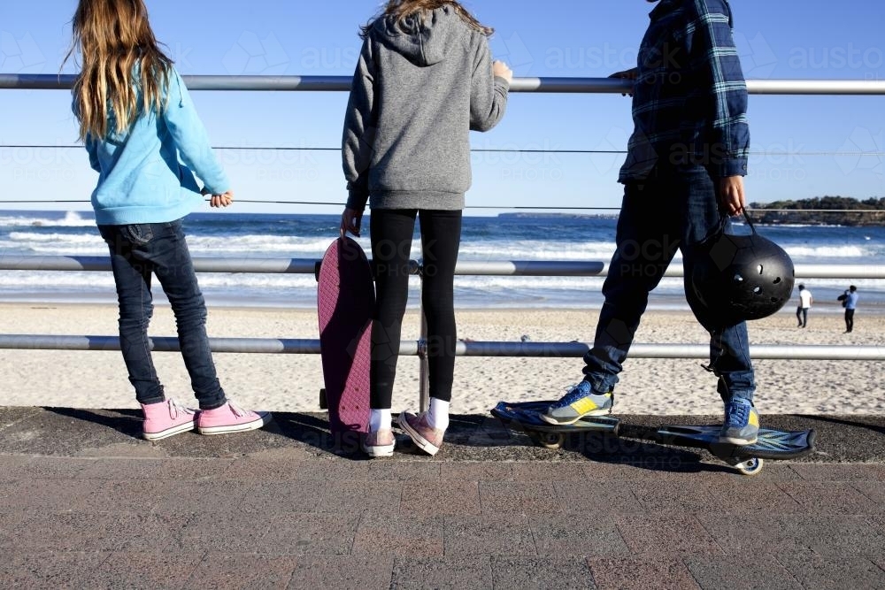 Three kids standing with skateboards looking out at the beach - Australian Stock Image