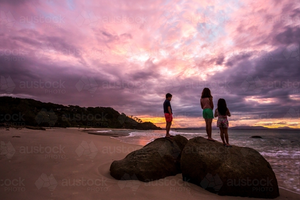 Three kids watching the sunset from a rock on the beach - Australian Stock Image