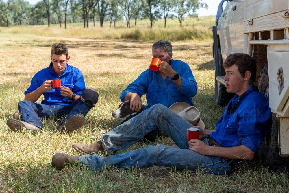 Three males sitting on ground next to ute having morning tea smoko. - Australian Stock Image