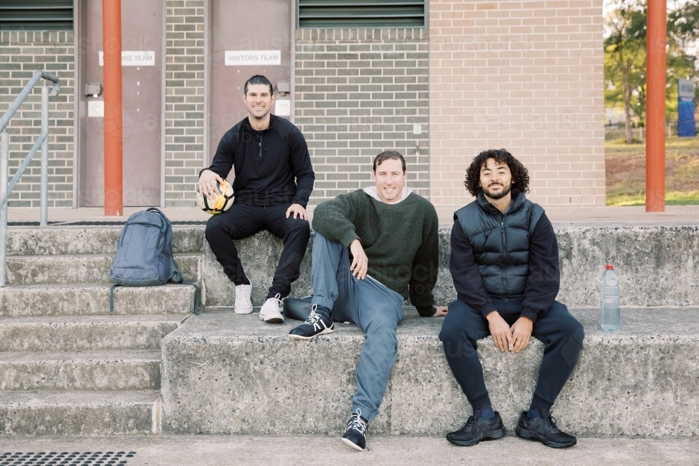 Three mates sitting on  soccer club steps, with one man holding a soccer ball - Australian Stock Image