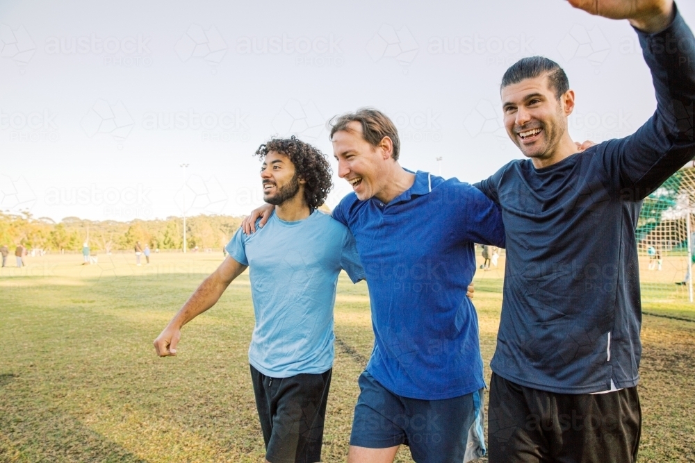 Three men walking together on a soccer field, laughing and smiling, celebrating a win - Australian Stock Image