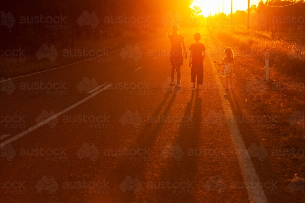 Three people walking along the roadside towards the sunset in golden light - Australian Stock Image