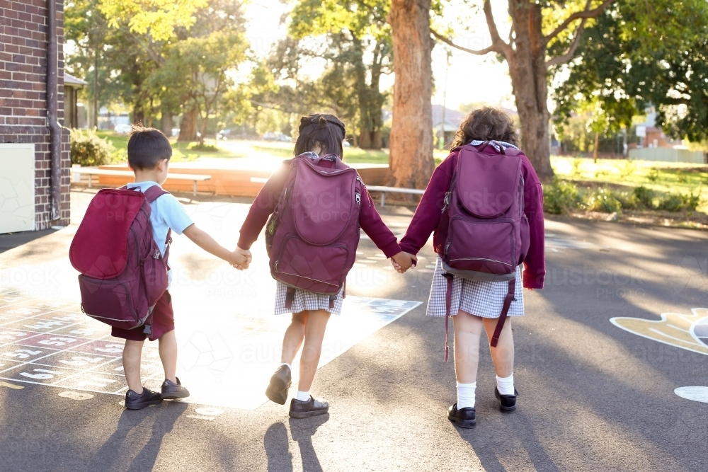 Three school children, holding hands, walking in school playground - Australian Stock Image