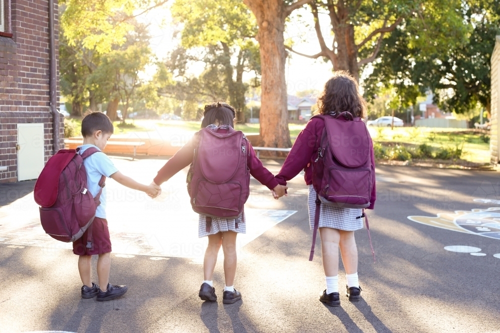 Three school children with backpacks on, walking through playground holding hands. - Australian Stock Image