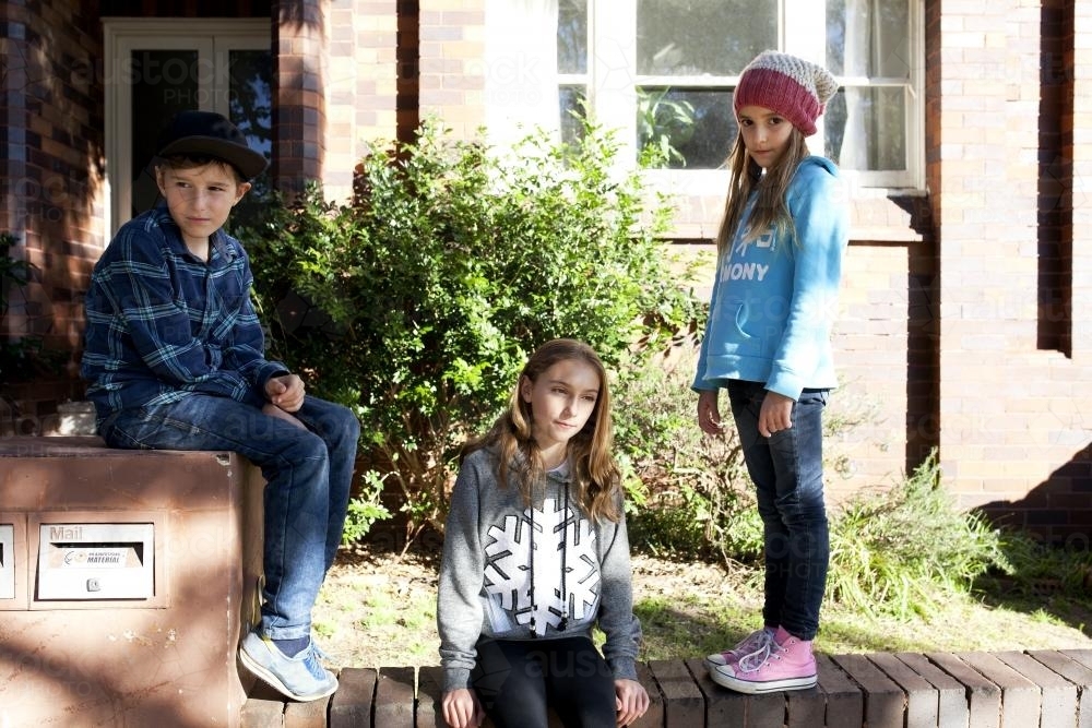 Three siblings posing on a brick fence in an urban front yard - Australian Stock Image
