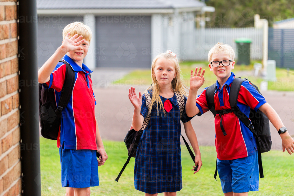 Three siblings ready for school waving goodbye at the door of home  - Australian Stock Image