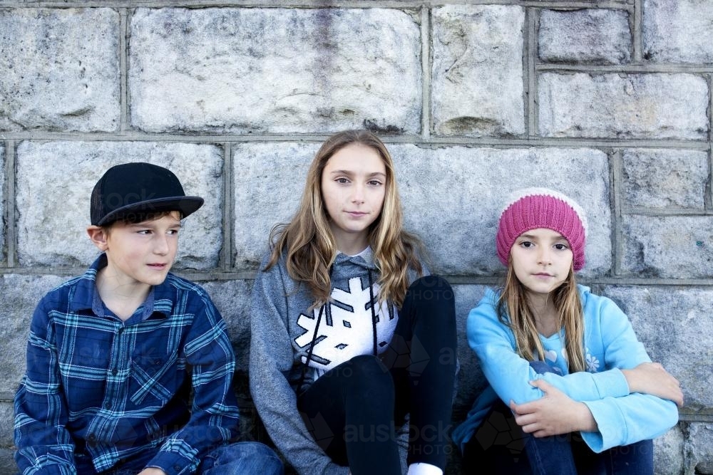 Three siblings sitting against a grey brick wall - Australian Stock Image