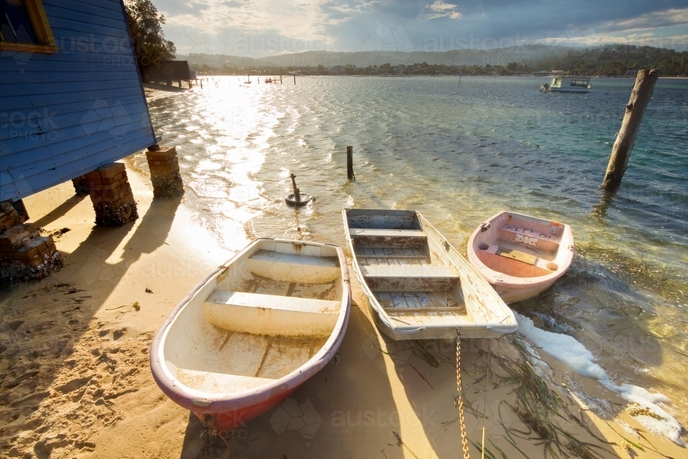 Three small rowboats tied together on a narrow sandy beach in the sunshine - Australian Stock Image