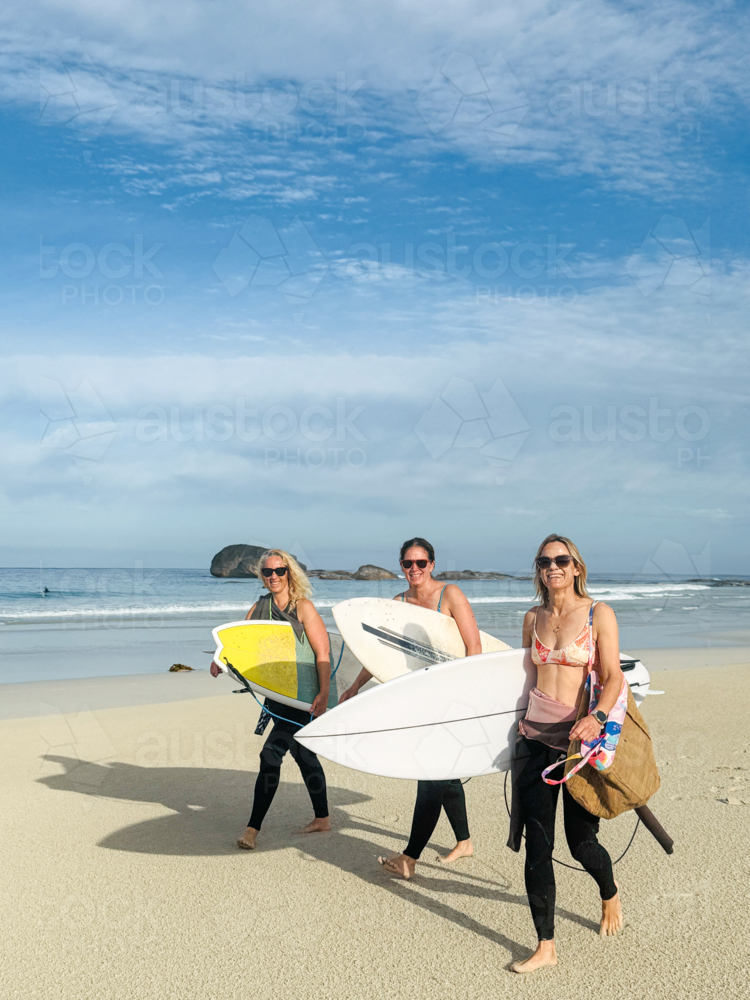Three surfer women walking along empty beach front carrying surfboards in warm morning light - Australian Stock Image