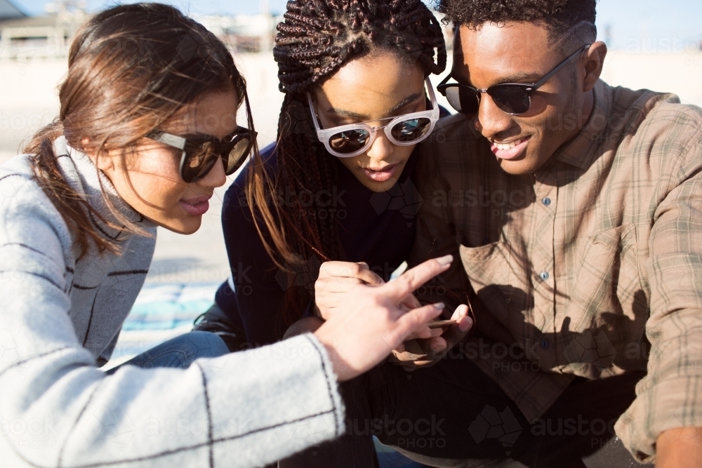 Three teenage friends sitting looking at a phone on a picnic rug - Australian Stock Image
