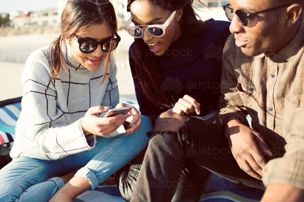 Three teenage friends sitting looking at a phone on a picnic rug - Australian Stock Image
