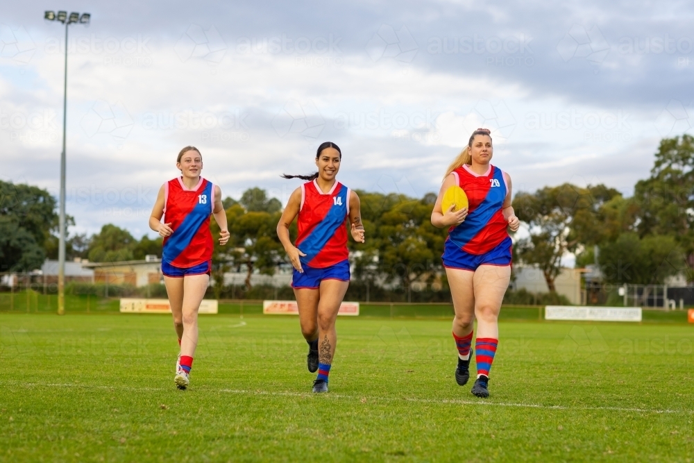 three women footballers jogging across football oval in red and blue uniforms - Australian Stock Image