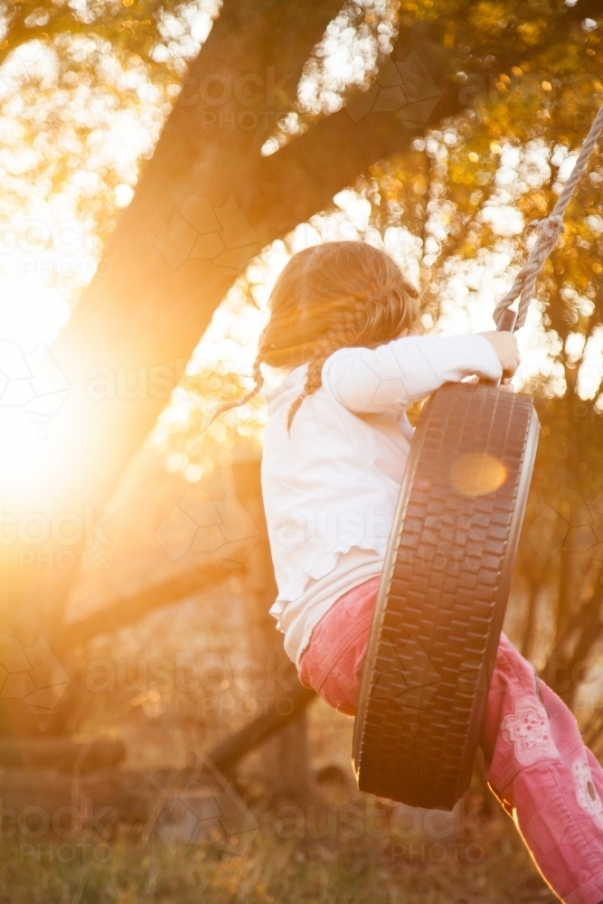 Three year old child swinging on tire swing with golden light sun flare - Australian Stock Image