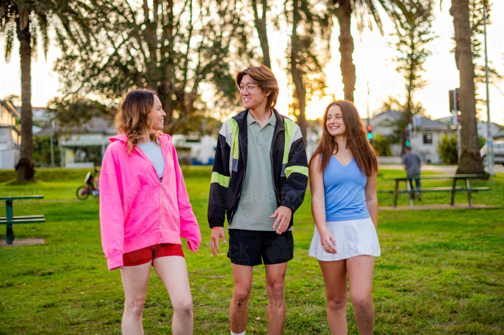 Three young adults leisurely stroll through a park as the sun sets behind palm trees - Australian Stock Image