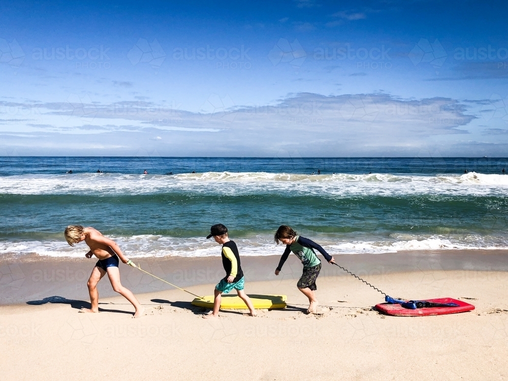 Three young boys dragging boogie boards along beach shoreline - Australian Stock Image