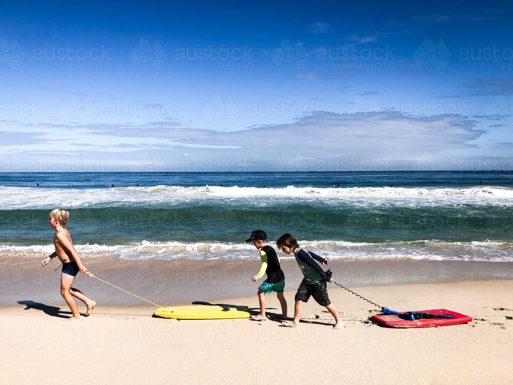 Three young boys dragging boogie boards along shoreline of beach - Australian Stock Image