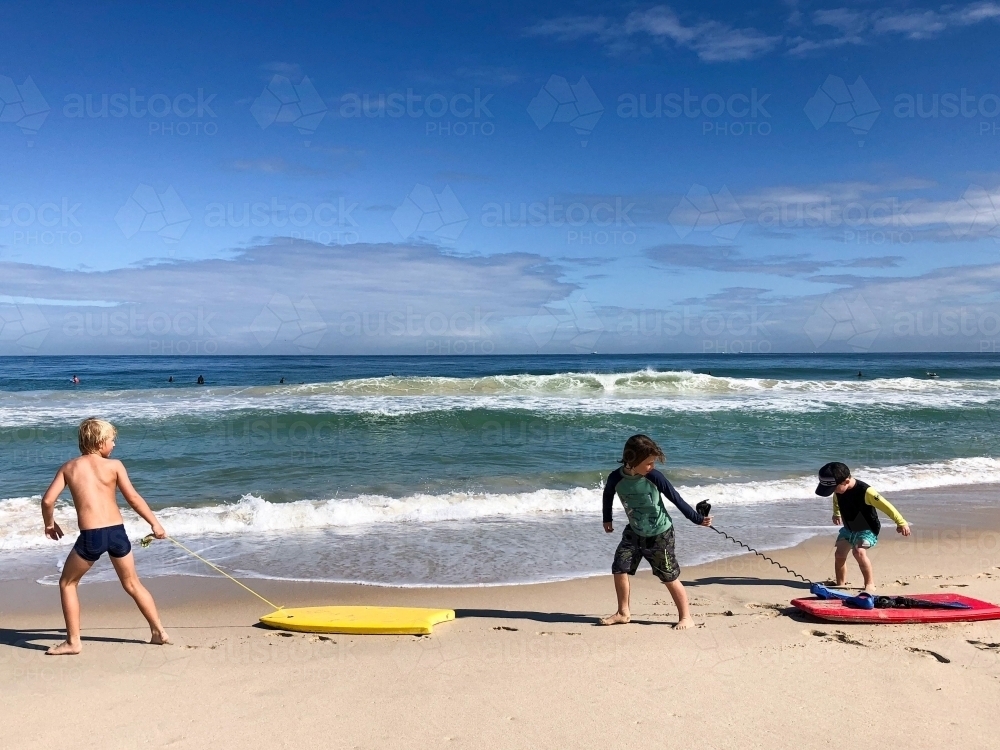 Three young boys dragging boogie boards along the shoreline of beach - Australian Stock Image