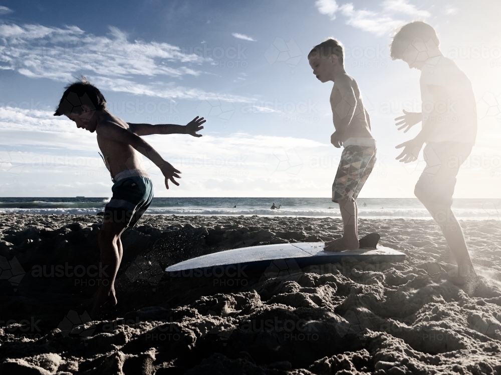 Three young boys playing on beach at sunset with surfboard - Australian Stock Image