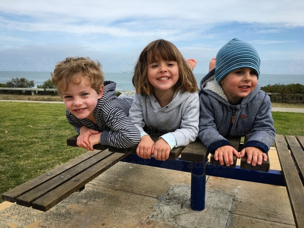 Three young children laying on park bench under overcast sky - Australian Stock Image