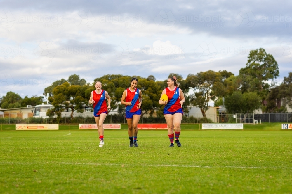 three young women wearing football uniforms running across oval - Australian Stock Image