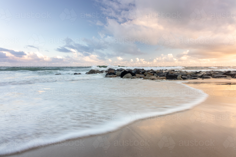 Tide coming in on beach at sunset with rock wall  in background - Australian Stock Image