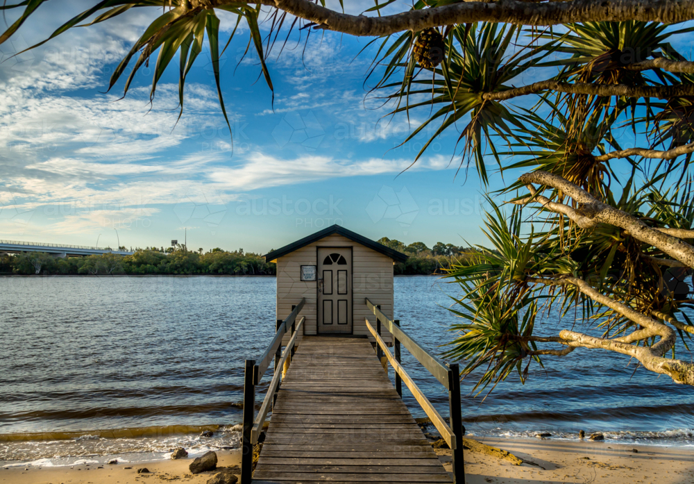 Timber boat shed and jetty on sandy river shoreline with pandanus tree, blue sky and clouds - Australian Stock Image