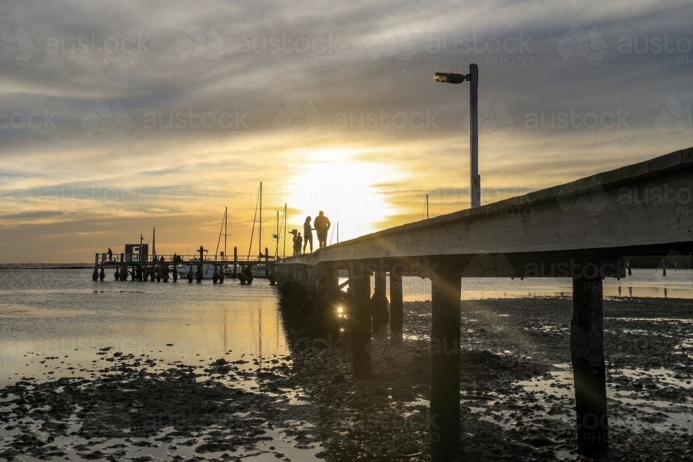 Timber jetty leading to sun setting through cloudy sky with people silhouettes - Australian Stock Image