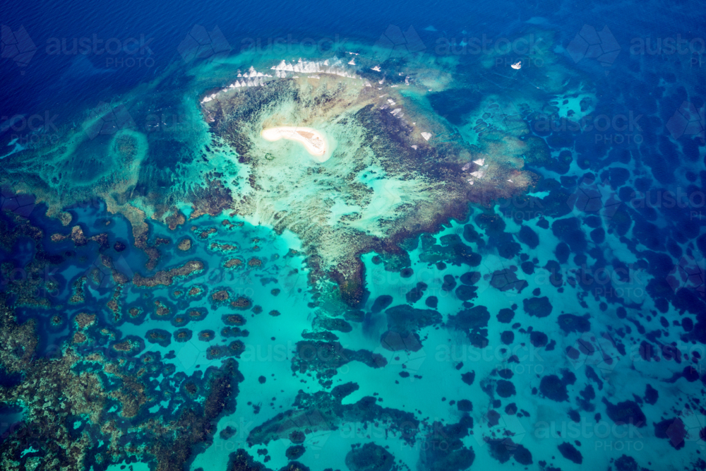 Tiny coral island surrounded by vivid turquoise reef and deep ocean blue. - Australian Stock Image