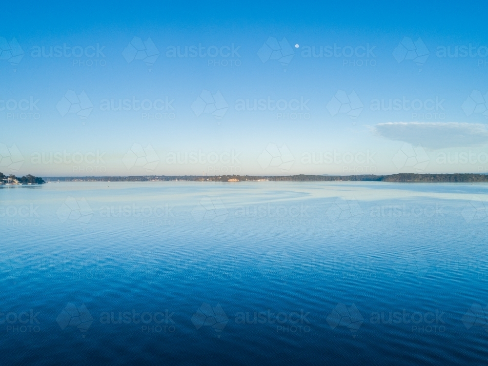 tiny moon in blue sky over softly rippling water of lake in Newcastle - Australian Stock Image