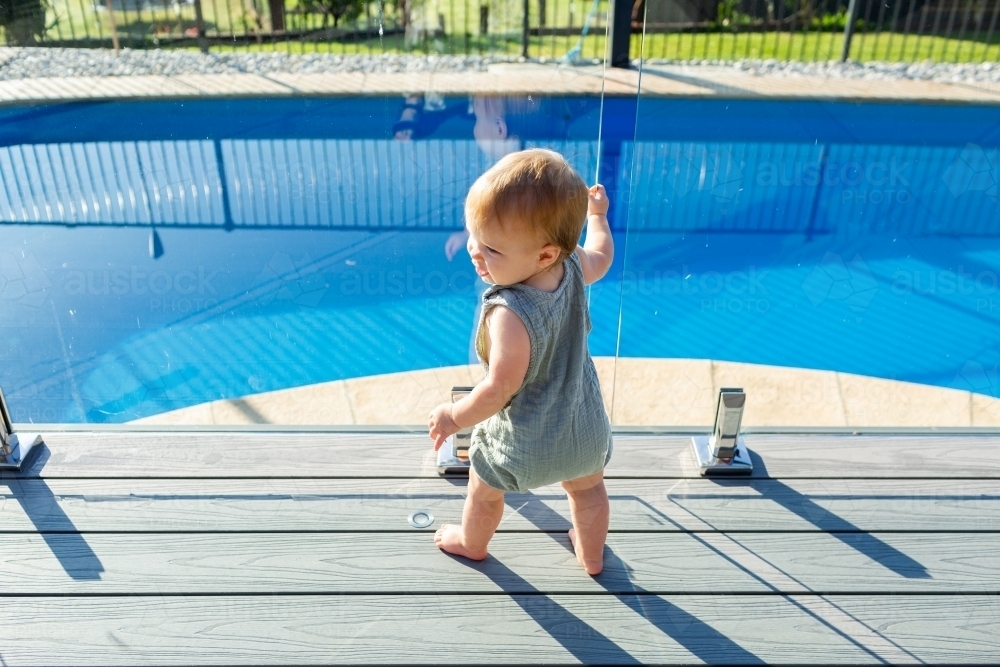 Toddler baby ten months old standing at glass pool fence beside inground backyard pool - Australian Stock Image