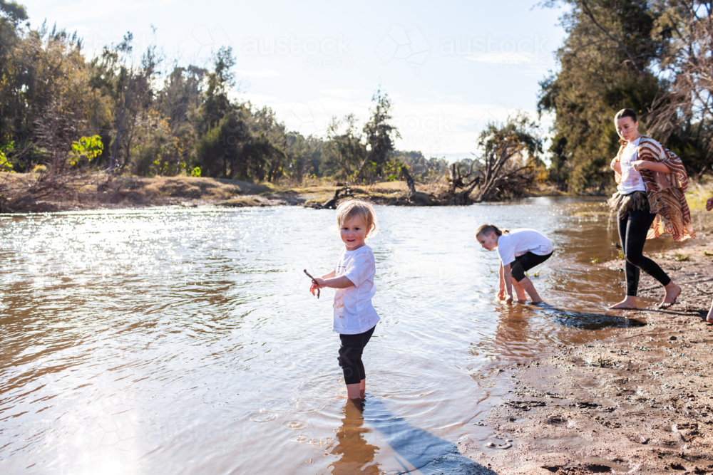 Toddler boy down by the river playing in water with siblings on bright sunlit morning - Australian Stock Image
