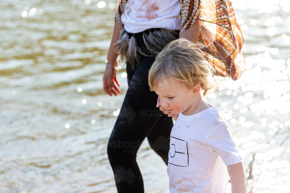 Toddler boy holding sisters hand while walking beside river in sparkling morning light - Australian Stock Image